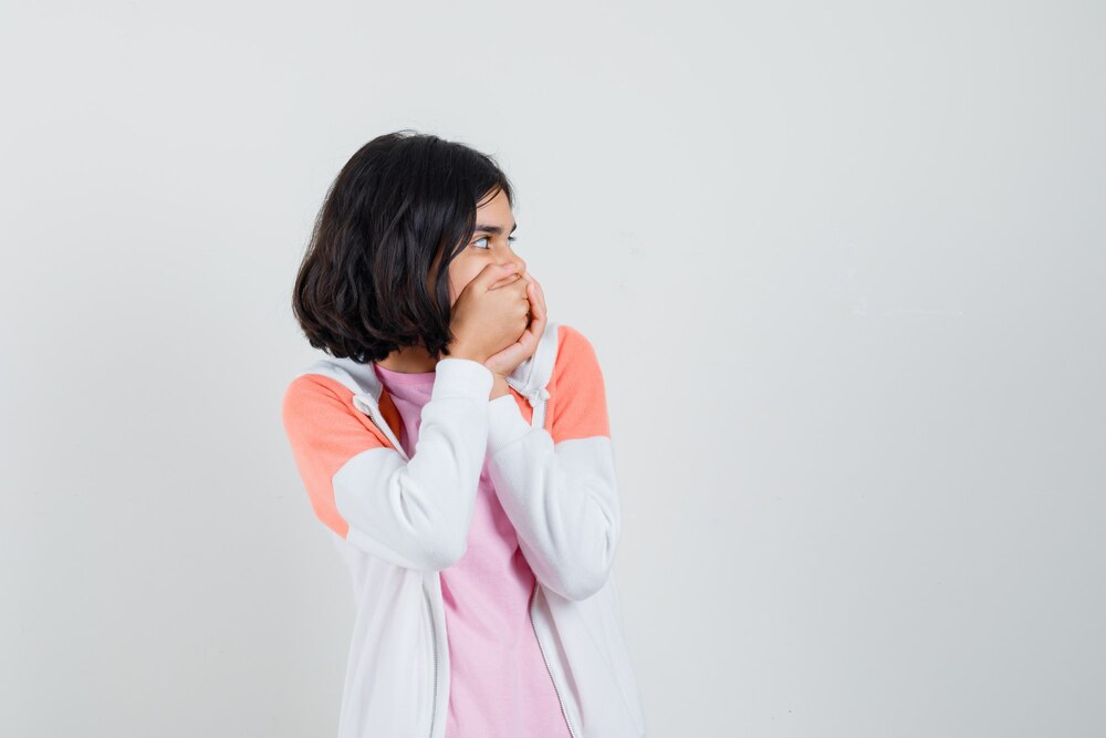 Young lady holding hands on her mouth in jacket, pink shirt and looking frightened.